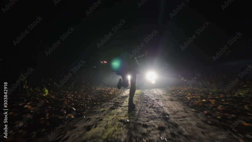 A frightened girl quickly runs away from a car following her along a country road at night. Silhouette of a running girl in the headlights of a car.