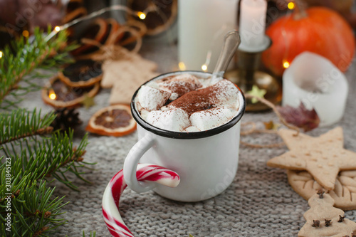 Cozy coffee break in a festive atmosphere. Mug of coffee or cocoa with marshmallows and Christmas cane among fir branches, candles, lights and holiday decor. Selective focus