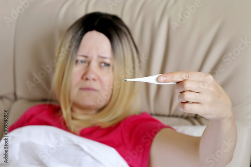 Sick woman lies in a bed with digital thermometer in hand, cold and flu season, selective focus. Concept of illness, fever, high body temperature