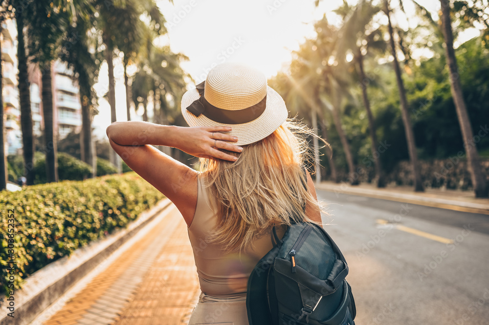 Chinese tourist walking in city streets on Asia vacation tourism. Caucasian beautiful woman in hat with backpack travel lifestyle concept. Walking street, green palms and highway in Sanya, Hainan. 