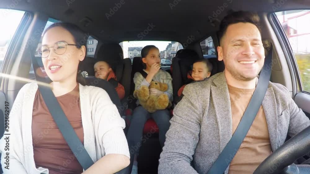 Action camera shot of cheerful man driving car and chatting with happy wife riding in passenger seat. Little girl and toddler twins sitting in backseat and snacking on crackers