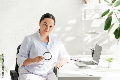 attractive dermatologist sitting at table and holding magnifying glass in clinic