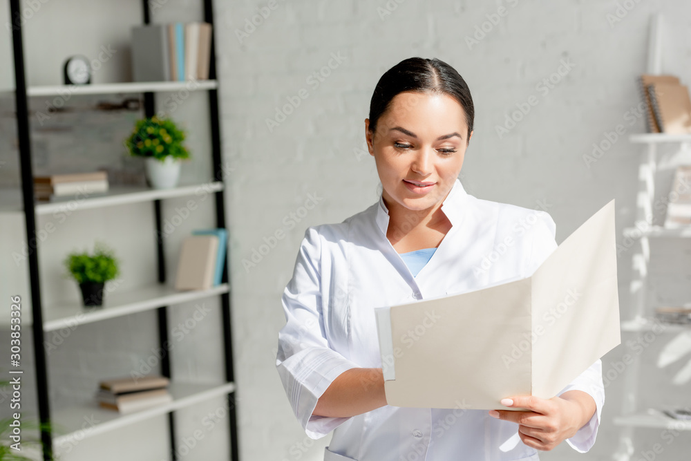attractive and smiling dermatologist in white coat holding folder in clinic