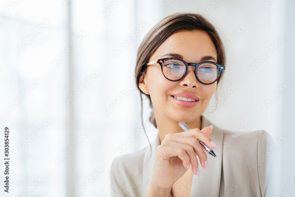 Smiling prosperous lady work in business sphere, holds pen, wears elegant clothes, has healthy skin, minimal makeup, looks somewhere, poses indoor against blurred white background, copy space left - obrazy, fototapety, plakaty 