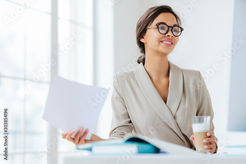 Cheerful businesswoman reads paper documents  focused aside with smile drinks milkshake sits at desktop makes business plan for project. Female executive manager in spectacles poses in coworking space