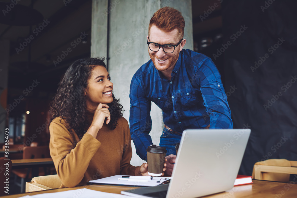 Fototapeta premium Positive multicultural male and female freelancers enjoying time for cooperating talking during coffee break near desk with laptop device, happy man and woman feeling excited from friendship
