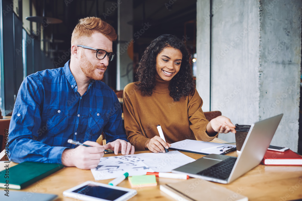 Successful graphic designers creating new logo for company during collaboration in coworking space, positive multicultural male and female hipsters enjoying time for cooperation on laptop device