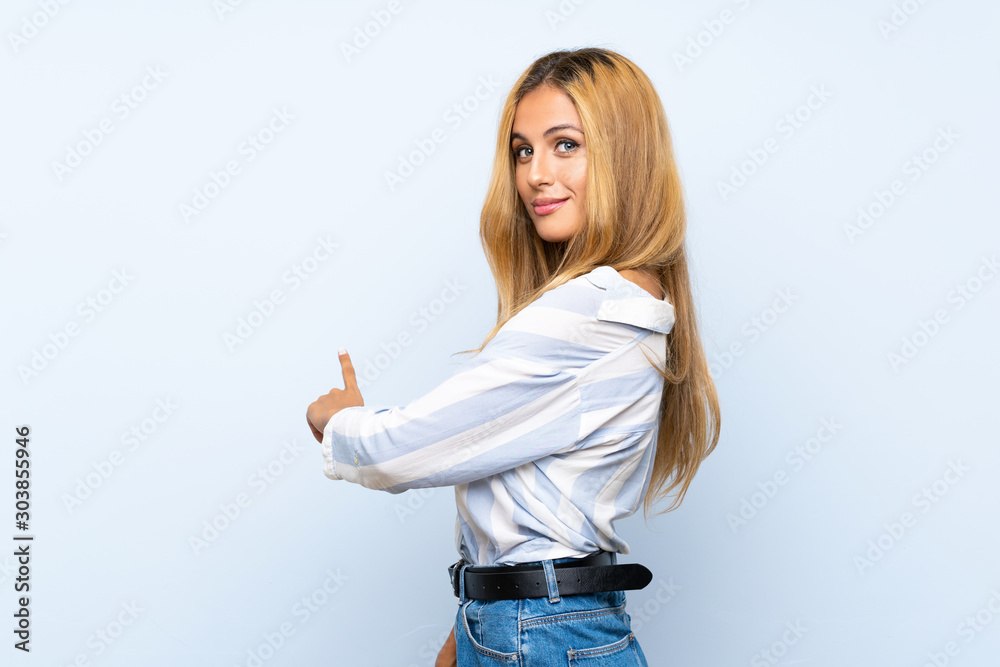 Young blonde woman over isolated blue background pointing back Young blonde woman over isolated blue background pointing back