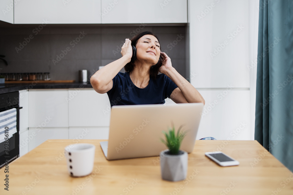Attractive and happy middle age female freelancer is working at her home. She is using headphones, listening to music and singing. Modern kitchen in background. Freelancing job concept. Attractive and happy middle age female freelancer is working at her home. She is using headphones, listening to music and singing. Modern kitchen in background. Freelancing job concept.