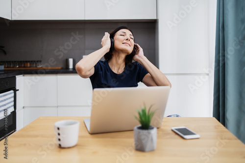 Attractive and happy middle age female freelancer is working at her home. She is using headphones, listening to music and singing. Modern kitchen in background. Freelancing job concept.