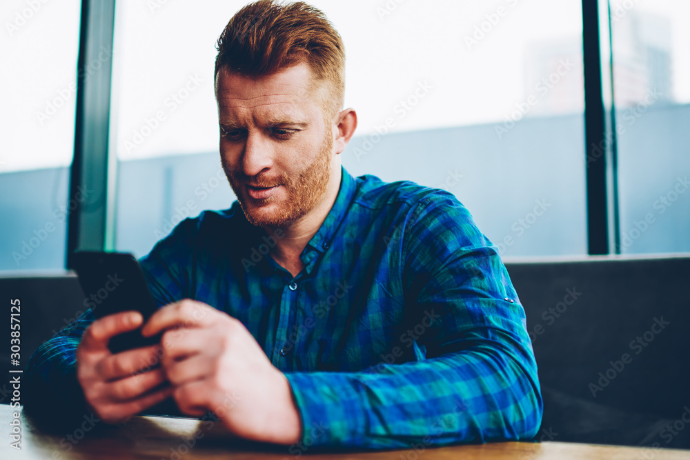 Male blogger with red hair charcking email and chatting online on smartphone connected to 4G internet sitting in coworking space.Young man in casual shirt reading incoming notification on cellular Male blogger with red hair charcking email and chatting online on smartphone connected to 4G internet sitting in coworking space.Young man in casual shirt reading incoming notification on cellular