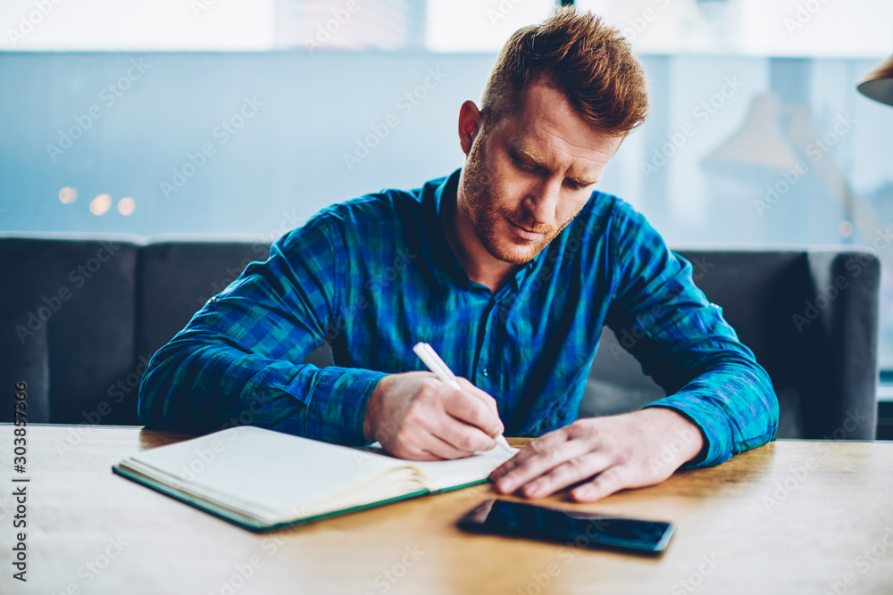 Skilled red haired student writing down homework in copybook studying at wooden table in coworking space.Pensive young man dressed in casual shirt noting checklist in notepad during free time - obrazy, fototapety, plakaty 