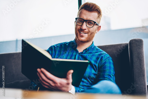 Portrait of cheerful handsome male entrepreneur with red hair smiling at camera while checking financial documentation sitting in own coffee shop.ositive student in eyeglasses with book in hands photo