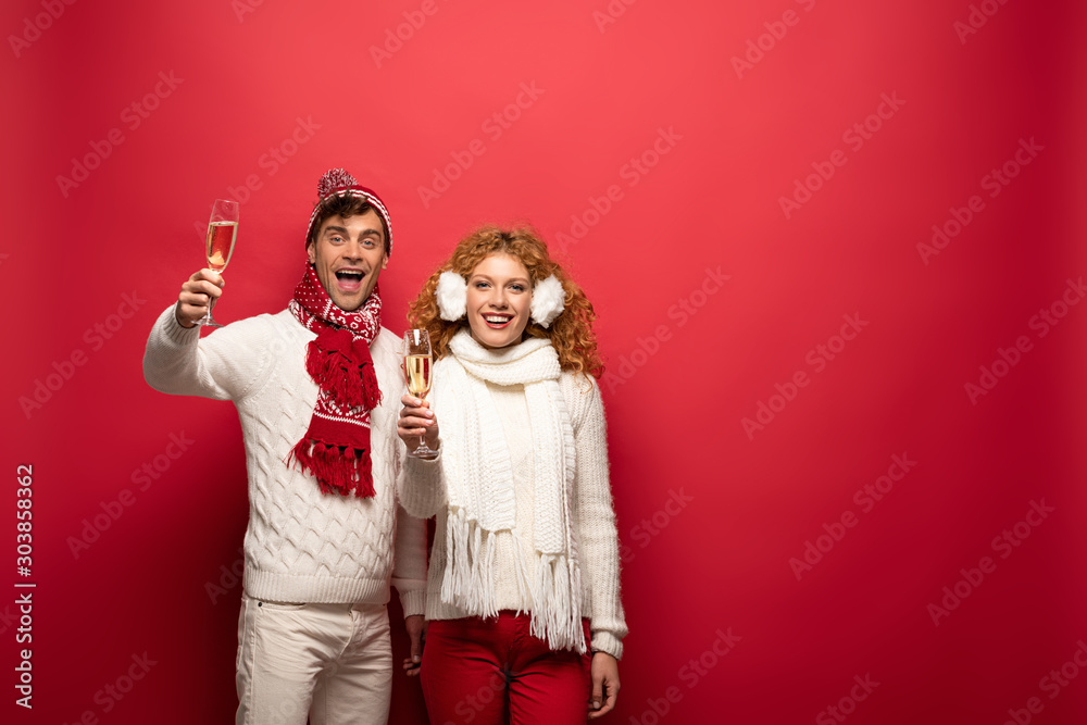 Fototapeta premium happy couple in winter outfit toasting with champagne glasses, isolated on red