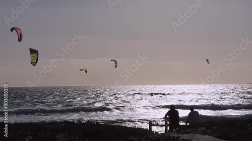 Timelapse of a Silhouette couple at beach watching kitesurfers catch big airtime and jumps. Windy summer’s day in Cape Town. Kiteboarding on glistening sea. Silver mirror ocean waves. Summer holiday 