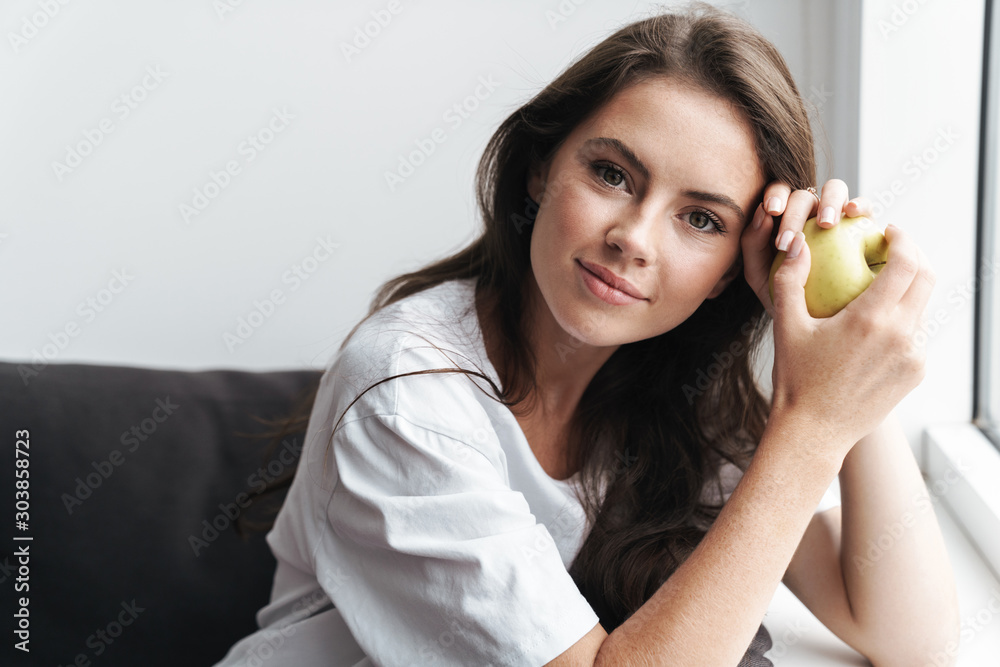 Image of young caucasian woman holding apple while sitting on sofa Image of young caucasian woman holding apple while sitting on sofa