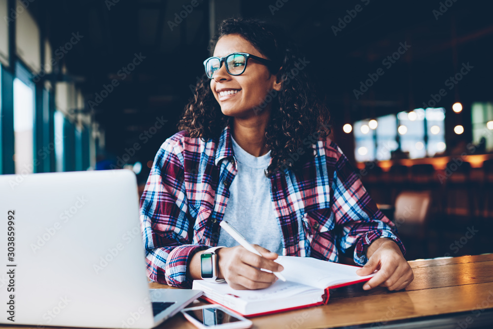Φωτογραφία Cheerful african american hipster girl in eyewear writing homework in notepad while looking away studying at laptop