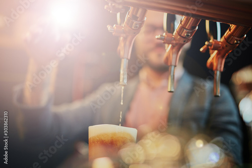 Tattooed caucasian barman pouring beer while standing in pub. Selective focus on glass.