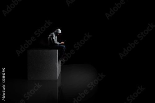  a lonely man with smartphone sits on a concrete cube in the dark. reflection on the floor.
