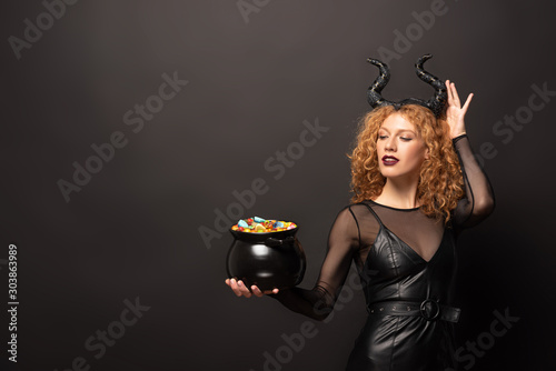 attractive woman in maleficent costume holding pot with candies for halloween on black photo