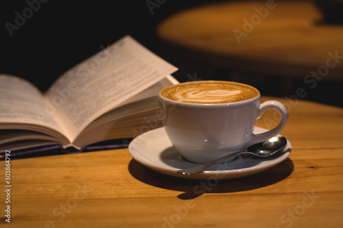 Ceramic cup of cappuccino in coffee shop with pattern on a wooden table with an open book. Latte art. Morning drink. Caffeine.