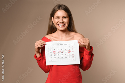 Young woman holding calendar with marked menstrual cycle days on beige background