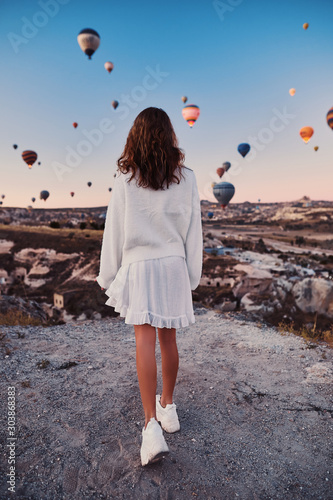 A tourist girl wearing white sweater on a mountain top enjoying wonderful view of balloons in Cappadocia