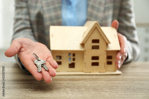 Real estate agent with house model and keys at wooden table, closeup