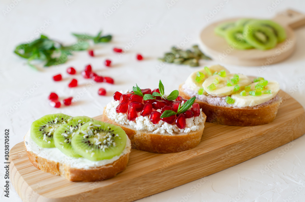 Open vegetarian sandwiches with vegetables, fruits and cream cheese on a wooden board on a white background. Kiwi and pomegranate in the background. Healthy breakfast concept. Copy space.