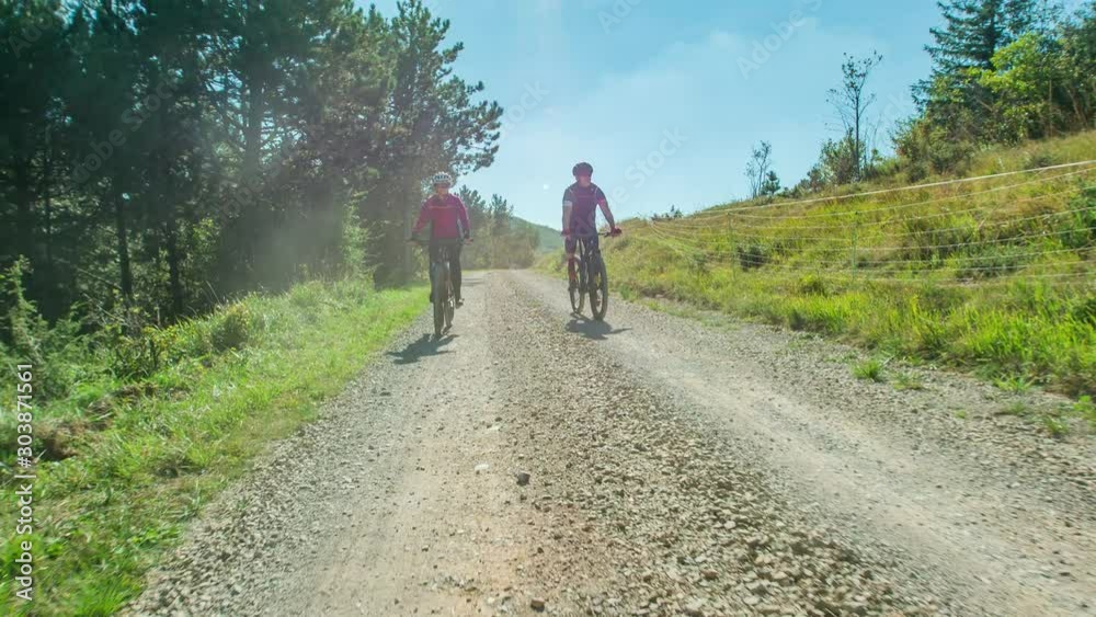 Pair cycle mountain bike down macadam road surrounded by grass and forest Pair cycle mountain bike down macadam road surrounded by grass and forest