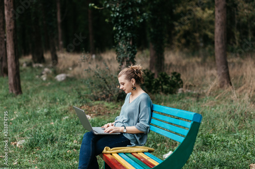 Fashionable young Caucasian woman sitting on colourful wooden bench in park. She is keyboarding on laptop. Freelance work concept. Copy space for text. Portrait. 
