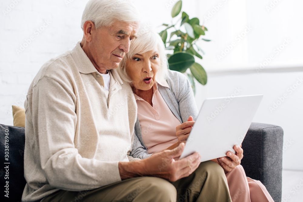 husband and shocked wife using laptop and sitting on sofa in apartment