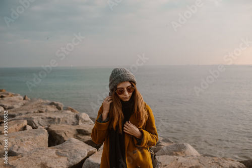 Girl portrait on the beach with the sea in the background. Stylish beautiful girl in mustard coat and glasses walking near the sea. 