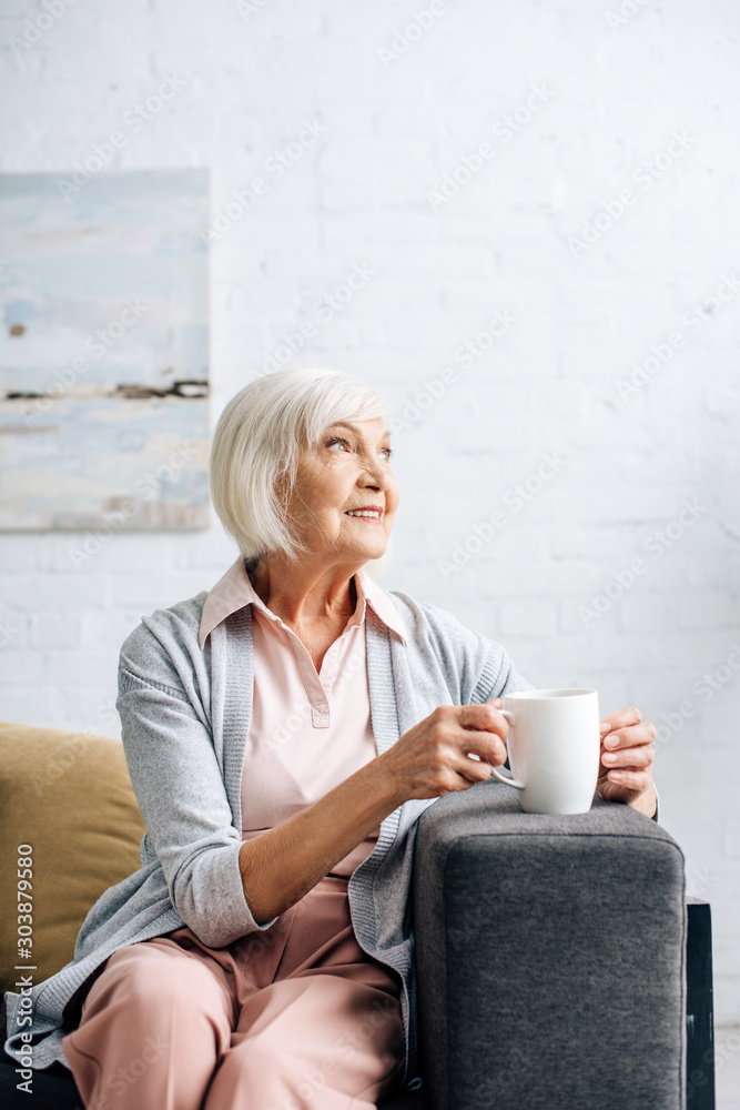 smiling senior woman sitting on sofa and holding cup in apartment smiling senior woman sitting on sofa and holding cup in apartment