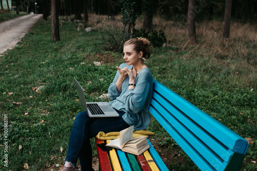 Fashionable young Caucasian woman sitting on colourful wooden bench in park. She is keyboarding on laptop. Freelance work concept. Copy space for text. Portrait. 