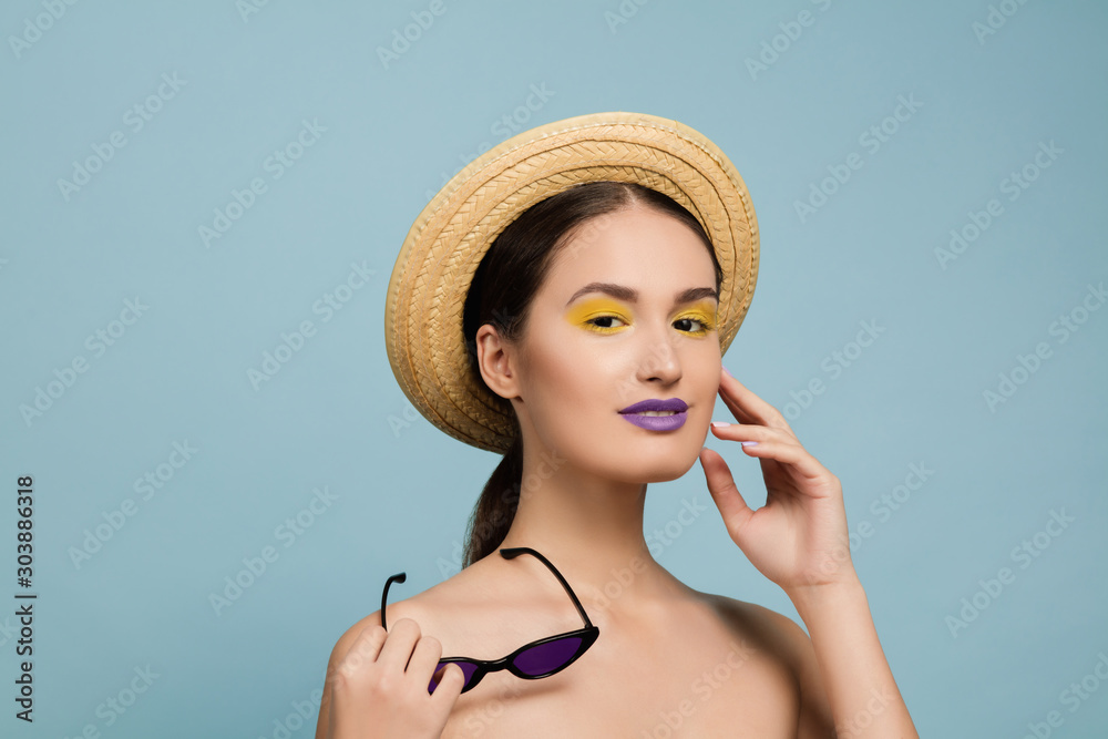 Portrait of beautiful woman with bright make-up, hat and sunglasses on blue studio background. Stylish and fashionable make and hairstyle. Colors of summer. Beauty, fashion, ad concept. Looks at