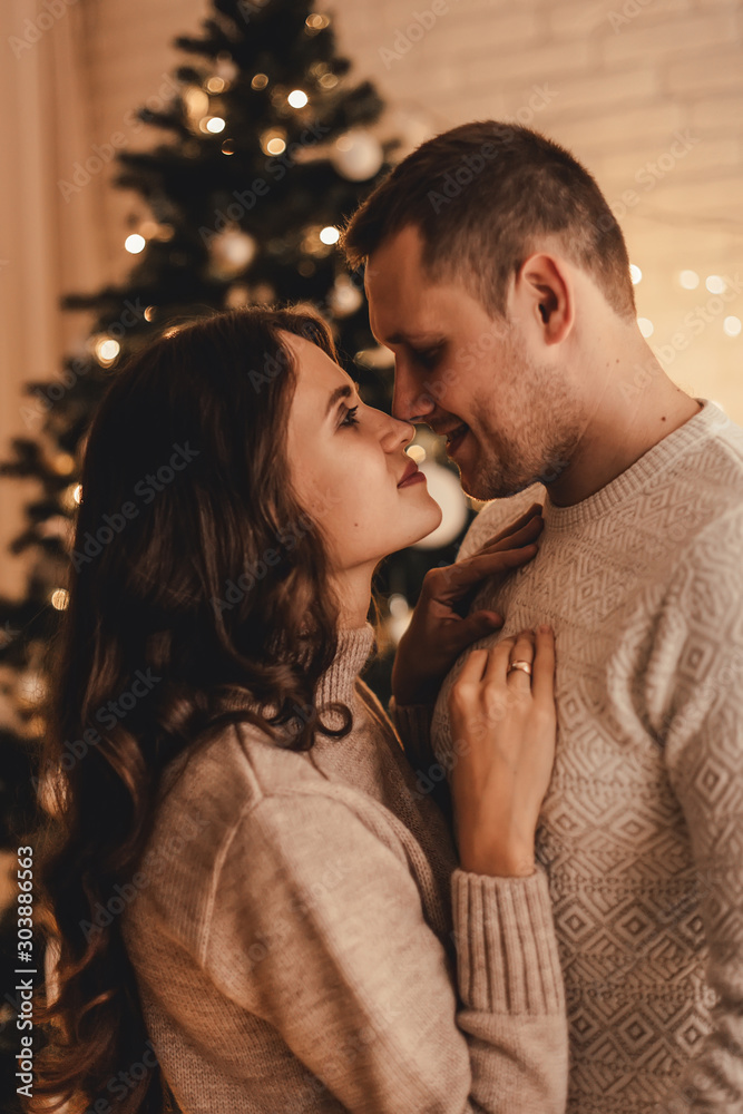 Lovely couple in sweaters hugging and celebrating new year in front of Christmas tree in decorated interior.