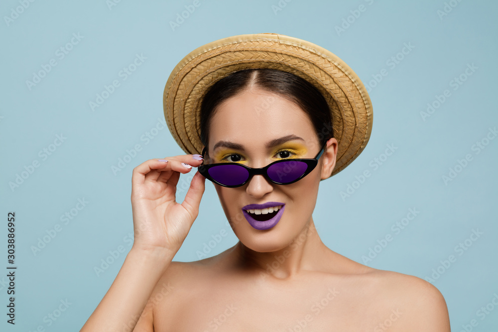 Portrait of beautiful woman with bright make-up, hat and sunglasses on blue studio background. Stylish and fashionable make and hairstyle. Colors of summer. Beauty, ad concept. Looks up of eyewear. Portrait of beautiful woman with bright make-up, hat and sunglasses on blue studio background. Stylish and fashionable make and hairstyle. Colors of summer. Beauty, ad concept. Looks up of eyewear.