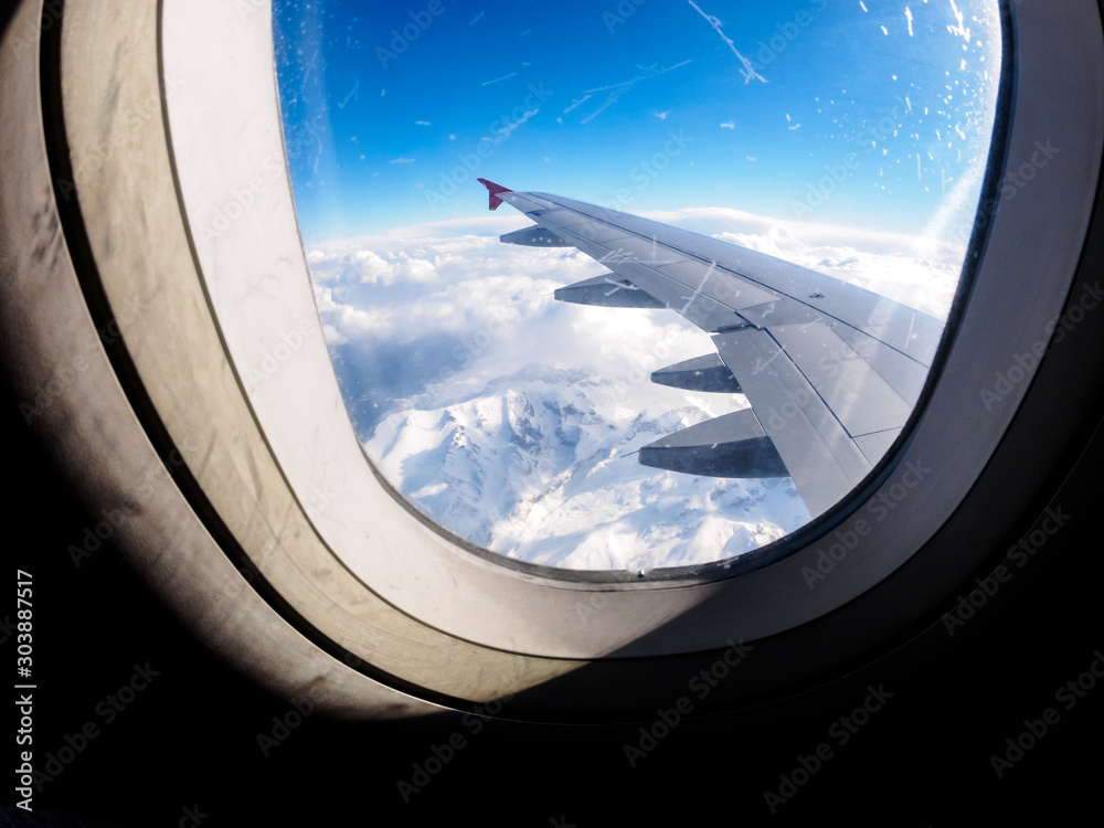 View of snow capped mountains from the airplane porthole. Sun shines through the window of the plane View of snow capped mountains from the airplane porthole. Sun shines through the window of the plane