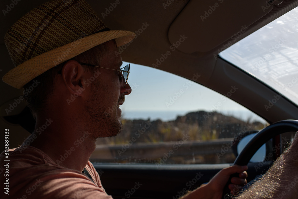 Handsome and smiling European man in sun hat and sunglasses is travelling by car along seaside. Insight view.