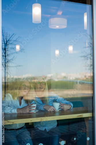 Two friends are enjoying coffee in a coffee shop  looking through a glass with reflections  sitting at a table chatting. Elderly Mother and adult daughter in a cafe. Mothers Day. Family resemblance.