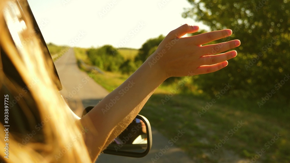 Girl with long hair is sitting in front seat of car, stretching her arm out window and catching glare of setting sun.