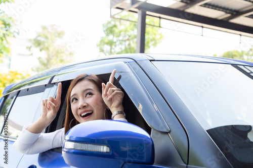 Happy Asian woman sitting in car and pointing up to empty copy space on top car roof © comzeal