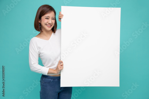 Smiling happy Asian woman holding and standing behind big white poster isolated on light green background