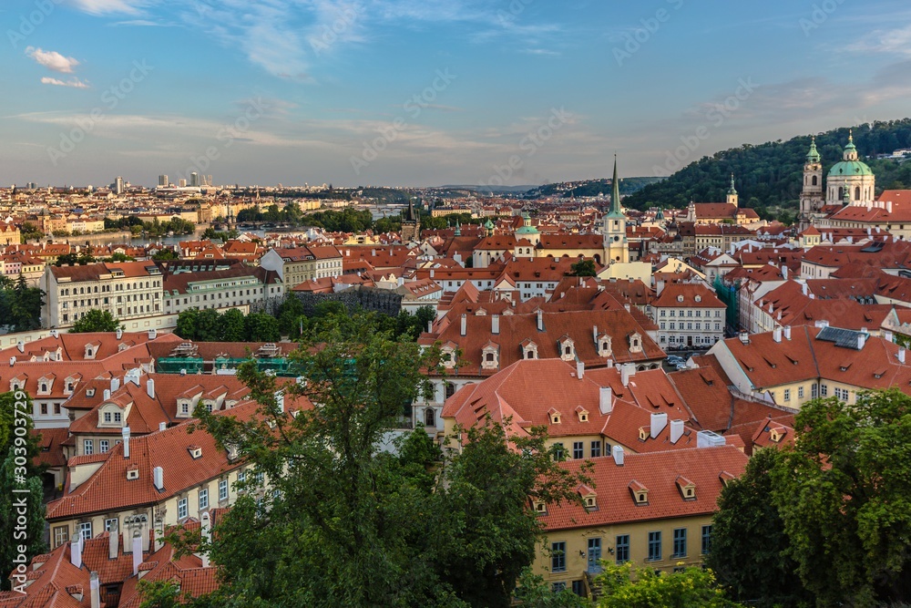 Prague, Czech Republic - July 20 2019: View of red roofs, cityscape with churches and historical buildings. Blue evening sky with clouds. Green trees in foreground.