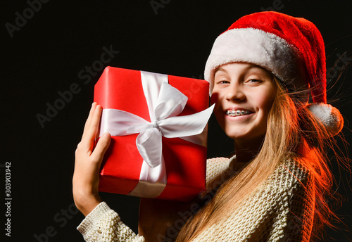 Happy smiling teenager girl in red cap with pompon with braces holding helong awaited new year gift red box with ribbon photo