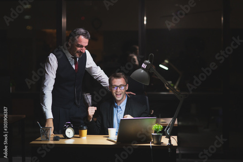 Caucasian boss working late stand with colleague in office at night. Business man look at laptop with coworker happy to success with job. Working late night and overtime concept
