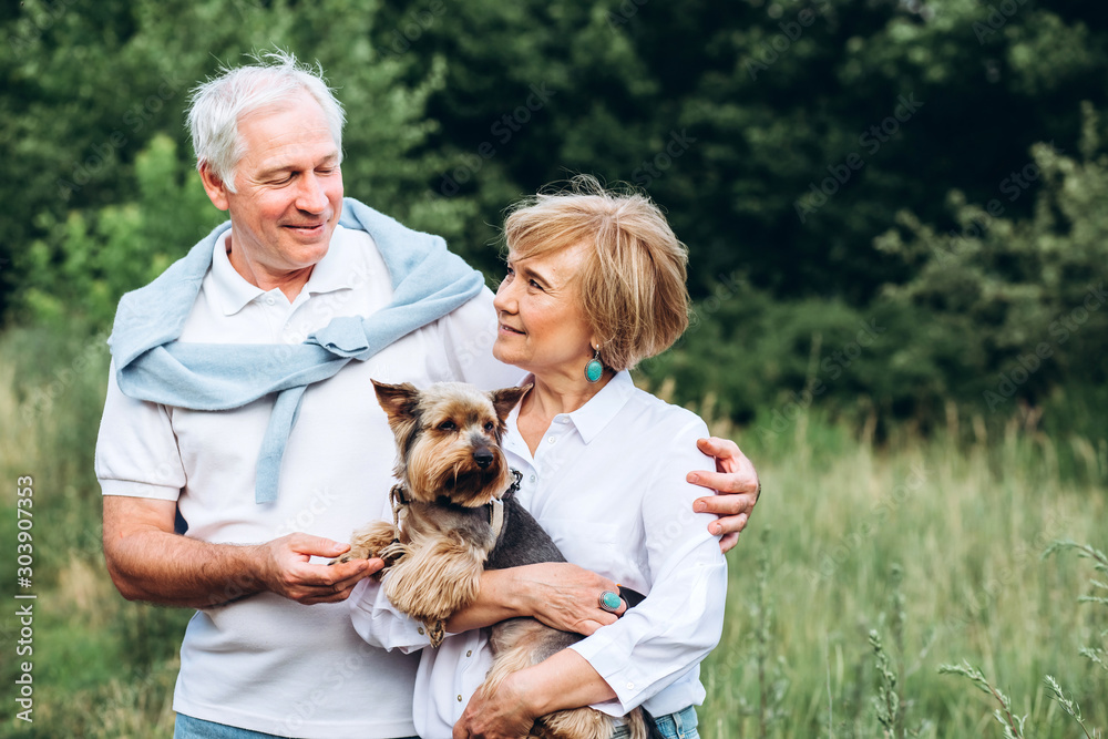 A mature couple is walking with a dog in a park. Elderly couple resting in nature with a dog. Close-up portrait of an elderly man and woman in white shirts and jeans. Stylish and modern grandparents. A mature couple is walking with a dog in a park. Elderly couple resting in nature with a dog. Close-up portrait of an elderly man and woman in white shirts and jeans. Stylish and modern grandparents.