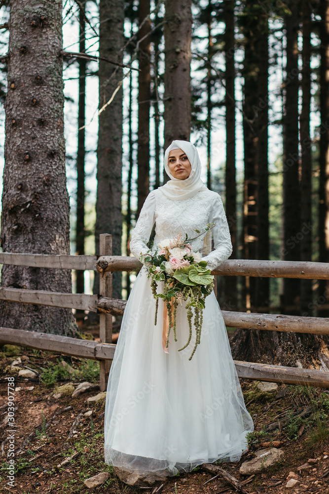 Fototapeta premium Beautiful elegant bride with hijab in lace wedding dress with long full skirt and long sleeves. She is holding a big bouquet of flowers. Outdoors, in the woods.