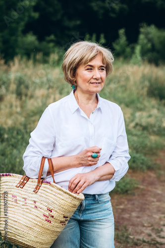 An elderly woman walks in the summer park. Close-up portrait of an old woman in white shirts and jeans. Stylish and modern grandmother with a wicker basket on the nature. © Stanislav